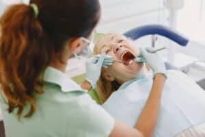 Dentist and assistant wearing gloves and masks examine a patient who is lying back in a dental chair with mouth open.