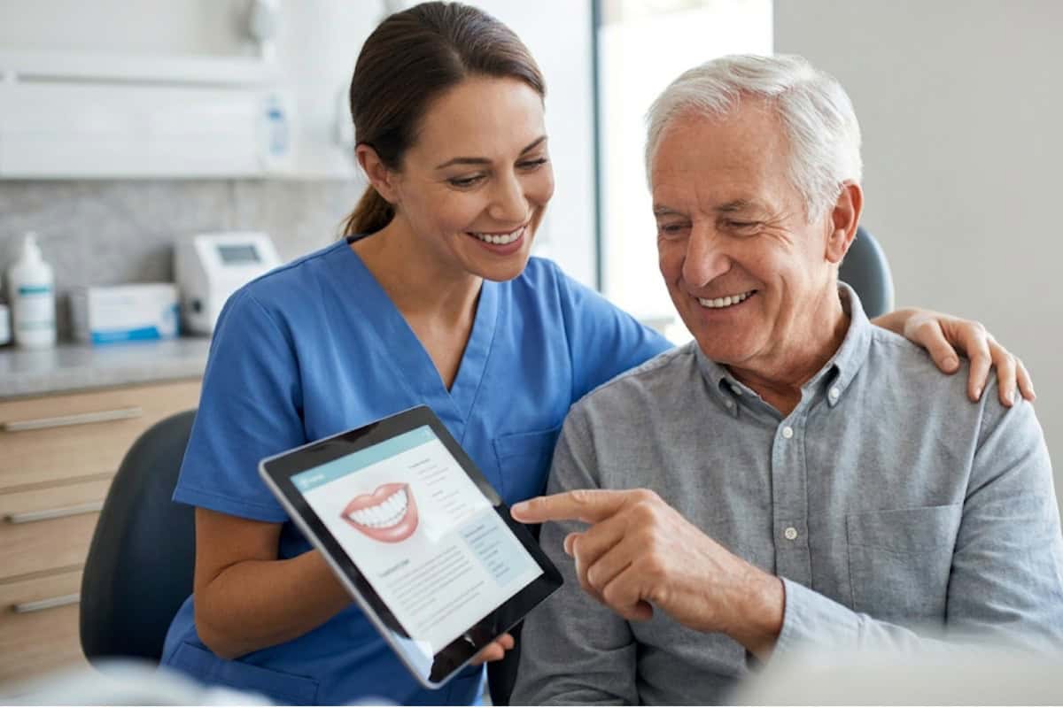 Dentist in blue scrubs shows an elderly man dental info on a tablet; both smiling in a clinic.