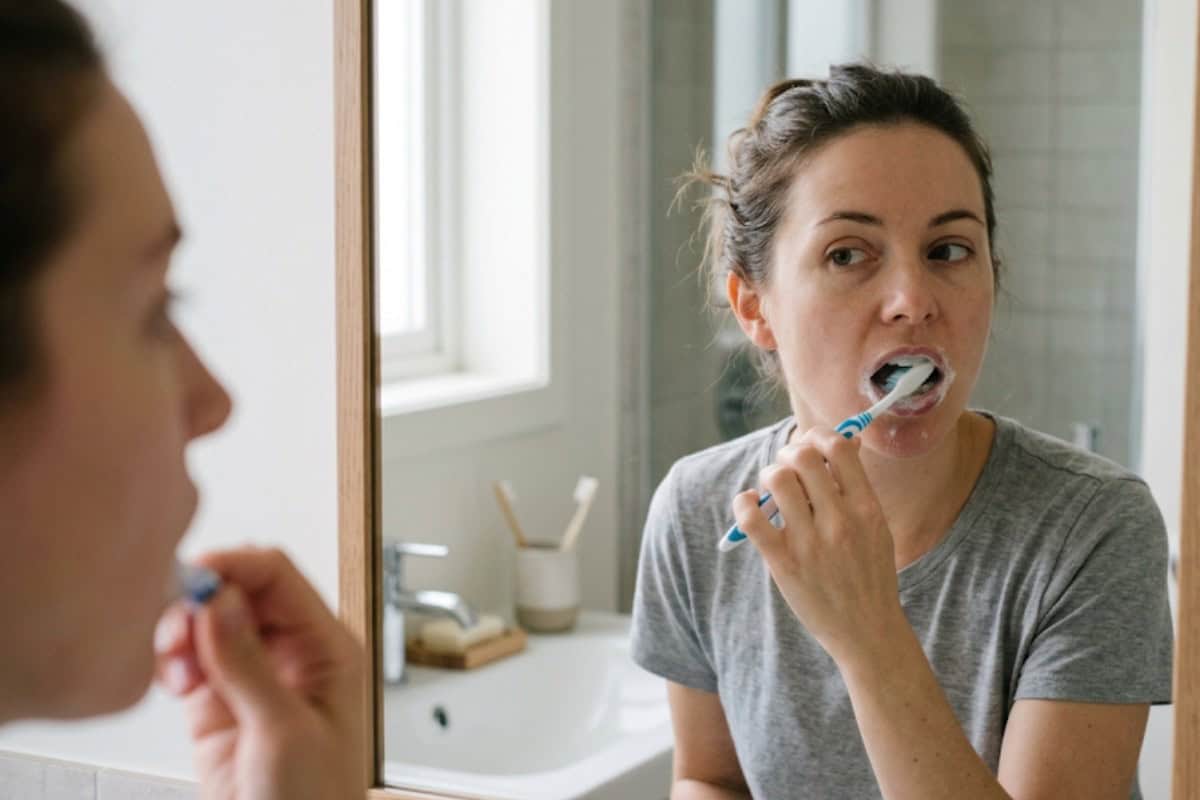 Woman brushing her teeth in a bathroom, looking at her reflection in a mirror with toothpaste foam around her mouth.