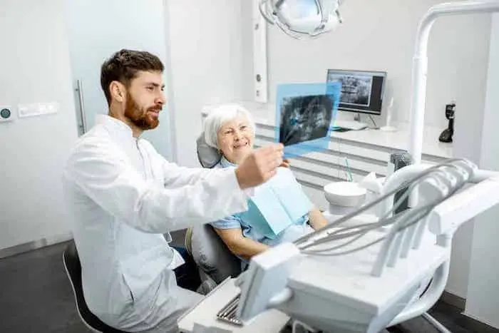 Dentist in a white coat showing an X-ray on a monitor to an older woman seated in the treatment chair.