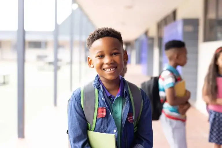 Smiling boy in a school corridor wearing a backpack, with other pupils behind him.