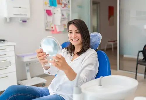 Woman seated in a dental chair holding a hand mirror and looking at her teeth.