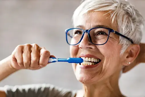 Older woman in glasses smiling as she brushes her teeth with a blue toothbrush.