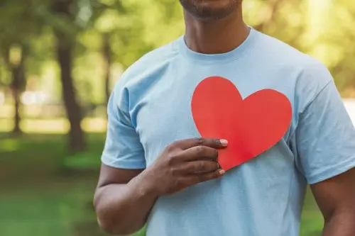 Man in a light blue T-shirt holding a red paper heart against his chest outdoors.