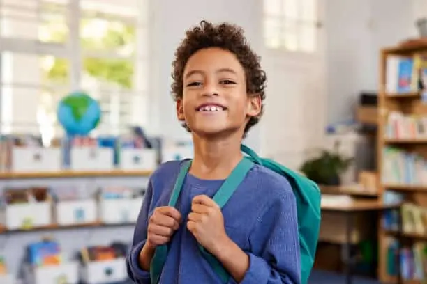 Boy in a classroom wearing a backpack and smiling.