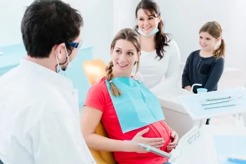 Pregnant woman seated in a dental chair with two clinicians and a child nearby.