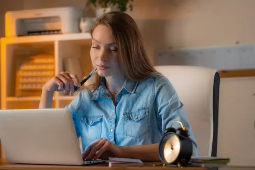 Woman at a desk working on a laptop with a pen to her mouth and a clock beside her.