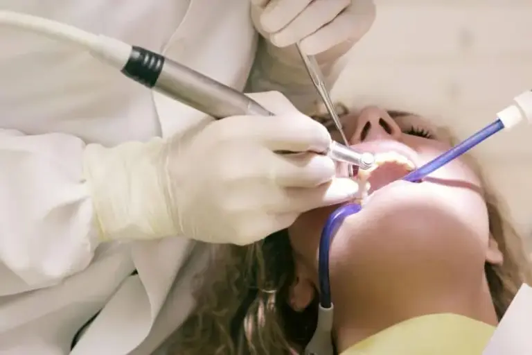 Gloved hands with a drill and suction working in a reclined patient's mouth.