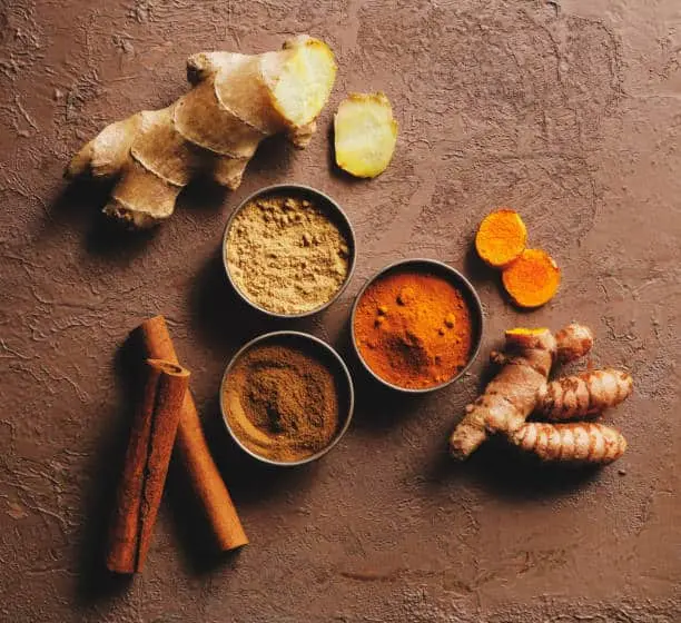 Bowls of ground spices with fresh ginger, turmeric root and cinnamon sticks.