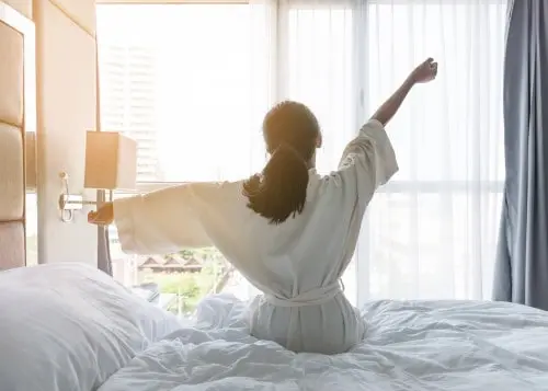 Woman stretching in bed in front of a bright window.