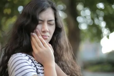 Young woman outdoors wincing with her hand pressed to her cheek.