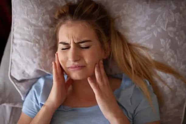Woman lying in bed holding both sides of her jaw.