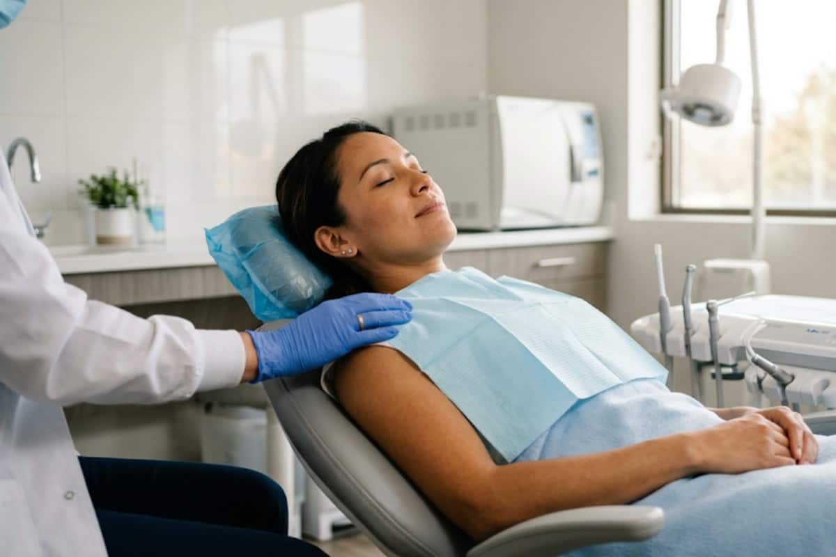 Woman reclining in a dental chair with eyes closed, wearing a blue bib, as a clinician in gloves rests a hand on her shoulder.